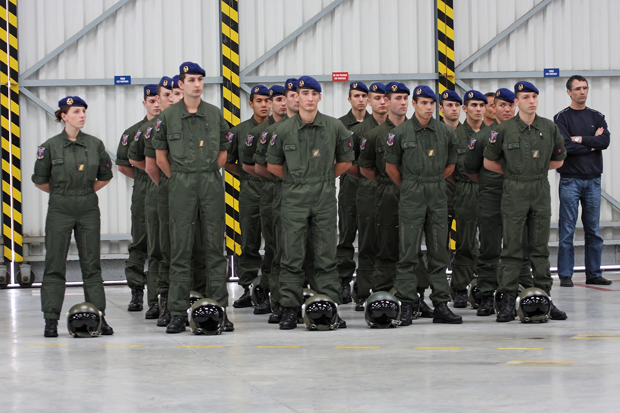 Élèves pilotes en combinaison de vol alignés dans un hangar, casque posé au sol, lors d'une cérémonie